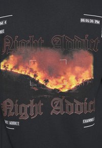 Svart t-shirt med en grafik av lågor på en kulle, med texten "Night Addict" i fet stiliserad font överlagd.