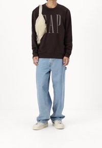 Tmavě hnědý svetr s logem "GAP", světlé modré volné džíny a béžové tenisky, doplněné béžovou crossbody taškou.
