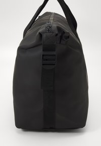 Sac de sport noir en matériau durable, doté d'une fermeture zippée, de poignées plates et d'accents en sangle. Texture lisse et mate.