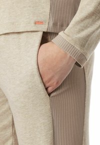 Beige Sportoberteil mit einem gerippten Seitenpanel und einer Seitentasche. Weicher Stoff mit subtiler Textur, ausgestattet mit einem kleinen rosa Etikett.