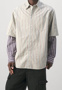 Mężczyzna ubrany w warstwowe koszule: krótki beżowy pasiak na długie rękawy w kratę fioletową i czarne spodnie cargo.