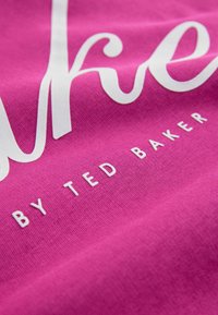 Il tessuto di cotone rosa presenta un ampio testo bianco con la parola corsiva "live" e, sotto, "by TED BAKER" in caratteri più piccoli. La texture appare morbida e liscia.