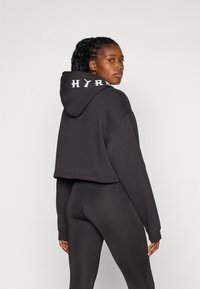 Beskyttet svart hettegenser med teksturert overflate og hvit "HYRD" lettering på hetten, kombinert med tettsittende svarte leggings.