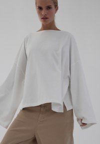 Biały, długi rękaw z dekoltem w łódkę, luźny krój, boczne rozcięcia i gładka tekstura, zestawiony z beżowymi spodniami.