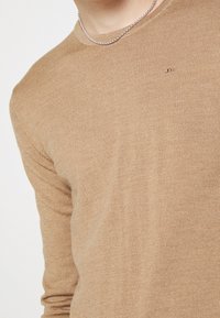 Pull beige en maille avec un col rond, présentant une texture subtile, des détails côtelés aux manches et un petit logo sur la poitrine.