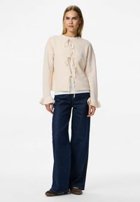 Suéter de punto rosa claro con escote redondeado, detalle de lazo y puños con volantes, combinado con pantalones de mezclilla oscuros de pierna ancha.