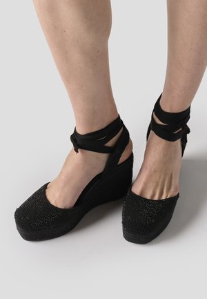 Stopala u crnim klinastim espadrilama s omotnim vezicama oko gležnja na svijetlosivoj pozadini.