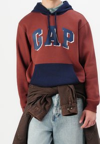 Luumu- ja laivastonsininen huppari, jossa valkoinen "GAP"-logo, edessä tasku, ja ruskea farkkupaita kerroksittain vyötärön ympärille solmittuna.