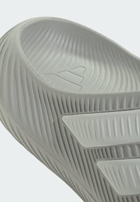 Primer plano de una sandalia tipo chancla gris con bordes texturizados y tres franjas blancas inclinadas en el lateral.