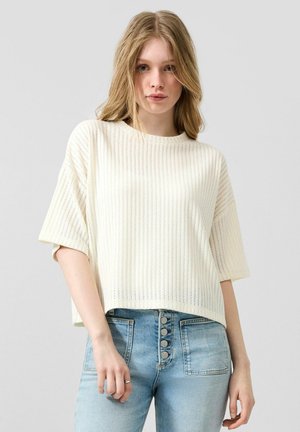 Jeune femme aux longs cheveux blonds portant un pull en maille blanc ample et un jean taille haute bleu clair avec des boutons visibles.