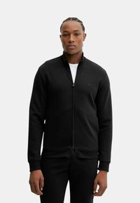 Svart zip-up sweatshirt med ståkrage, ribbat midja och ärmslut, tillverkad av mjukt material. Har en subtil logotyp på bröstet.