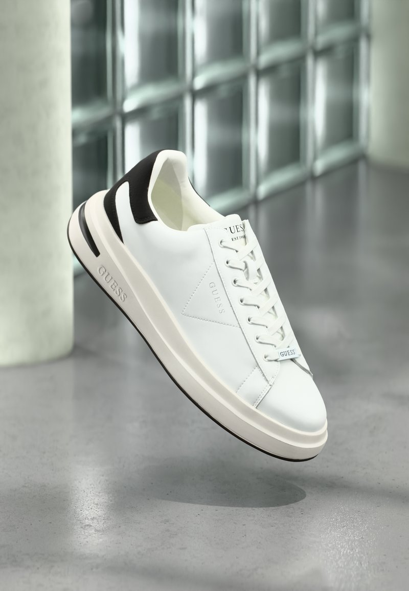 Sneakers bianchi in pelle con accenti neri, chiusura a lacci piatta e suola texture. Presentano il logo "GUESS" sul lato e sulla linguetta.