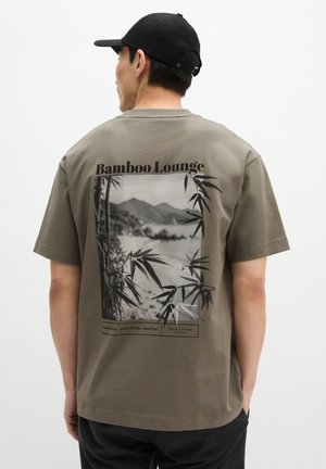 Mann trägt schwarze Kappe und olivgrünes T-Shirt mit dem Schriftzug "Bamboo Lounge" sowie Bambus- und Bergstrand-Grafik am Rücken.