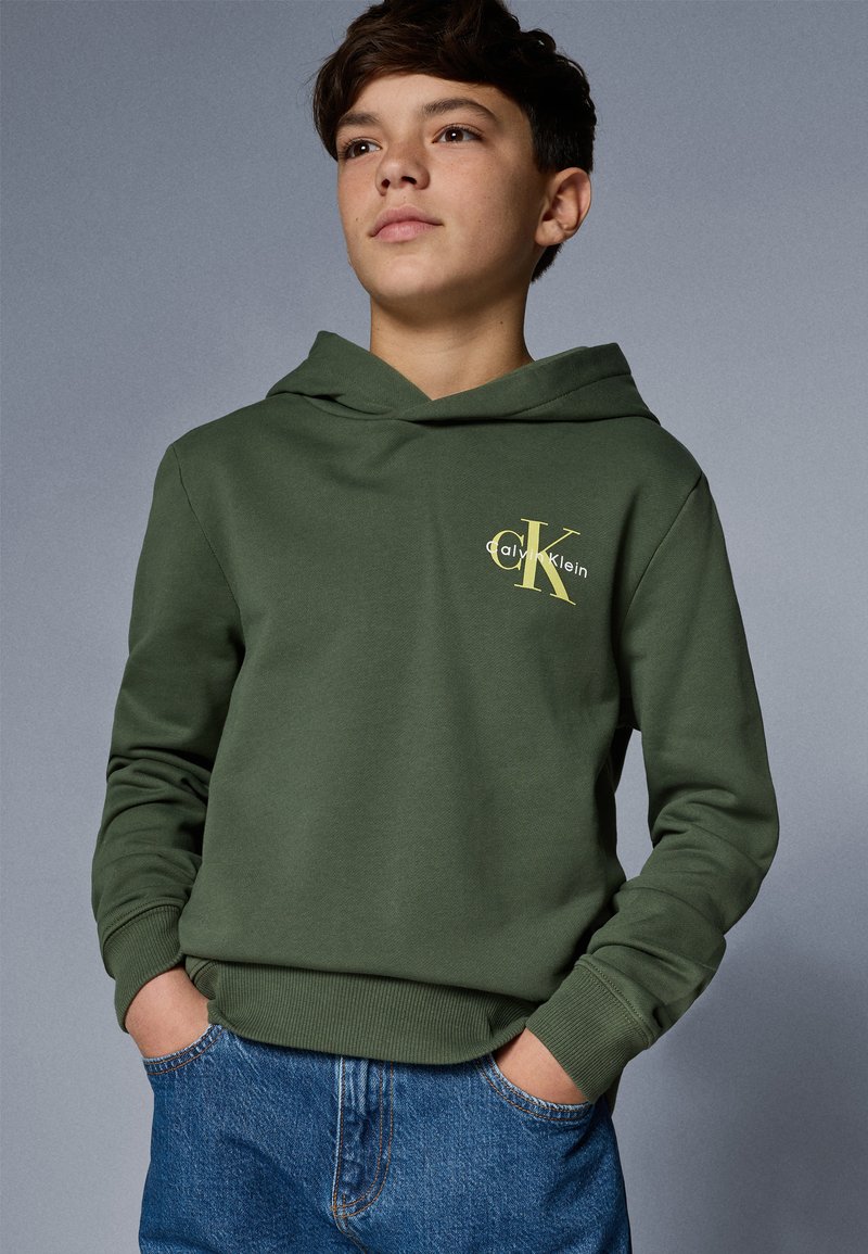 Grüne Kapuzenjacke mit gelbem Calvin Klein Logo auf der Brust, mit gerippten Bündchen und Saum, kombiniert mit blauen Jeans.