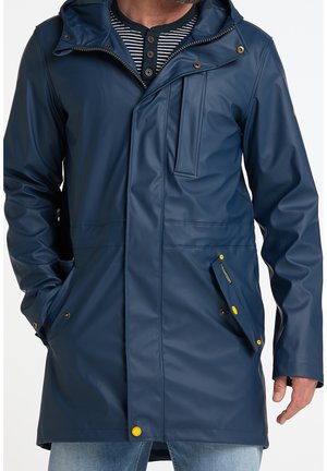 Homme portant un long imperméable à capuche bleu marine avec des boutons-pression jaunes et les mains dans les poches sur un jean bleu clair.