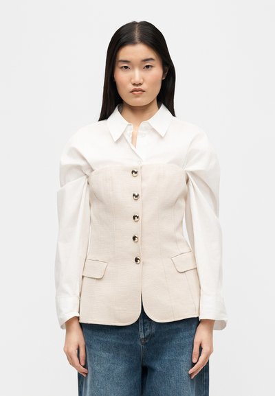 Γυναίκα με μακριά σκούρα μαλλιά φοράει λευκό πουκάμισο κάτω από κορσέ μπεζ με κουμπιά και μπλε τζιν, στέκεται μπροστά από μονόχρωμο φόντο.