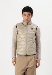 Gilet smanicato beige imbottito con motivo trapuntato, zip nera e piccolo logo. Indossato sopra una maglia a maniche lunghe con texture.
