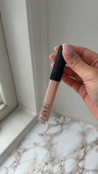 Main tenant un gloss à lèvres beige NARS avec un capuchon noir au-dessus d'une surface en marbre près d'une fenêtre.