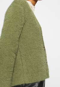 Cardigan verde in maglia fuzzy con bottoni indossato sopra una maglietta in pizzo bianco e pantaloni neri, mostrato di profilo su una persona.