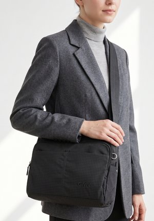 Femme portant un blazer gris et un col roulé, porte un sac bandoulière noir avec un motif quadrillé et le logo « GMV » à l'avant.