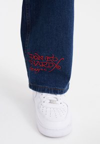 Mörkblå denimjeans med röd broderad "Don Ed Hardy designs"-logotyp vid fållen, kombinerat med vita sneakers med perforerade detaljer.
