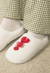 Weiße, plüschige Hausschuhe mit einem bestickten roten Hummerdesign oben. Getragen über beigen Socken, kombiniert mit grün-weiß karierten Hosen.