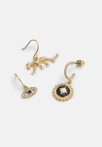 Brincos em tom dourado com um design de leopardo, um motivo de olho com acentos de cristal, e uma peça circular com uma pedra preta, todos variando em formas.