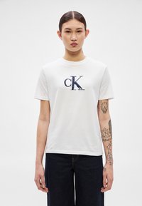 Valge puuvillane T-särk, mille rinnal on tumesinise tikandiga "ck" logo, ümar kaelus ja lühikesed varrukad. Kandmisega tumedad teksad.