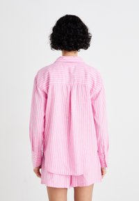 Camisa oversized azul e branca com listras, punhos com botão, pregas nas costas e bainha reta; tecido macio; mostrada de costas.