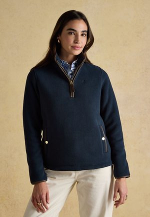 Woman wearing navy quarter-zip fleece over blue striped shirt and cream pants, standing against beige textured background.
