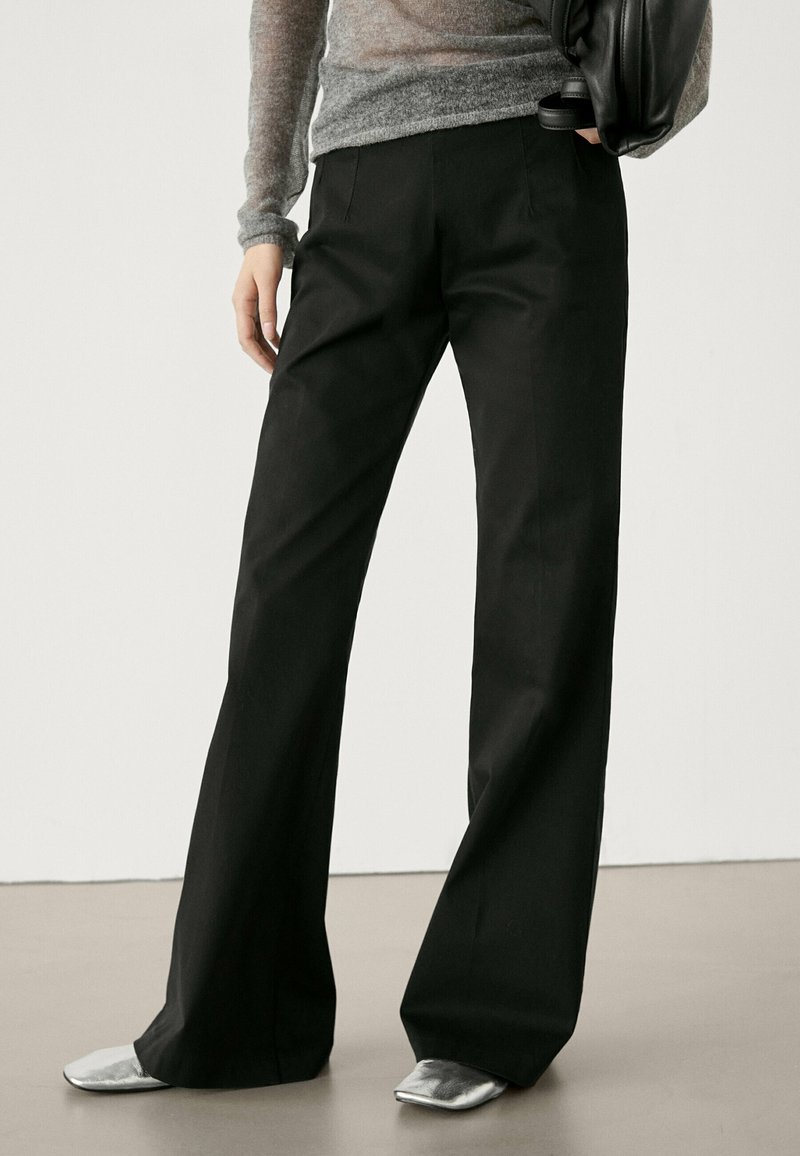 Personne portant un pantalon large noir, un haut à manches longues gris et des chaussures argentées à bout carré, tenant un sac noir au niveau de la taille.