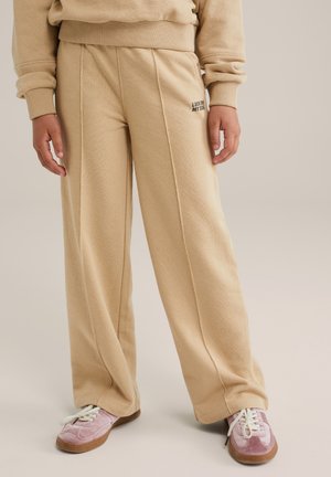Personne portant un pantalon beige à jambes larges avec des détails de couture à l'avant et un petit texte noir, associé à des baskets roses et blanches sur un fond clair.