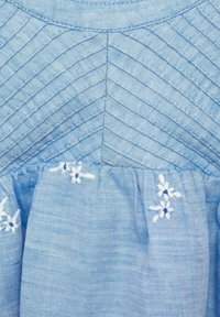 Helles blaues Gewebe mit Zickzack-Steppnähten an der Oberseite und kleinen weißen gestickten Blumen nahe der gerafften Naht darunter.