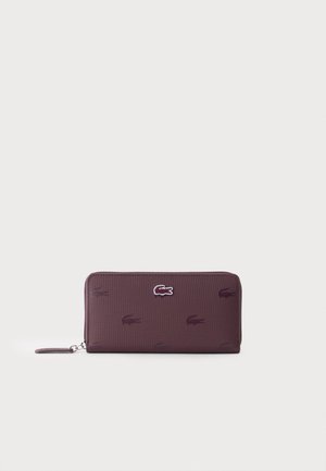 Portefeuille zippé violet foncé avec surface texturée et logos crocodile répétés, un en métal argenté, centré sur le devant.