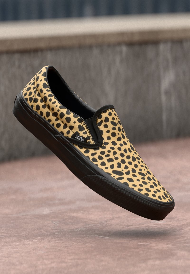 Baskets à enfiler avec motif tacheté jaune et noir et semelle noire, flottant au-dessus d'une surface pavée.