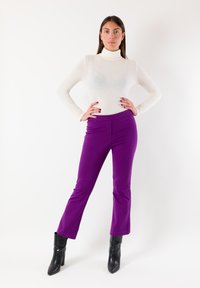 Maglia bianca a collo alto abbinata a pantaloni viola a vita alta e svasati. La modella indossa stivali neri con tacco. Texture lisce in tutta l'immagine.