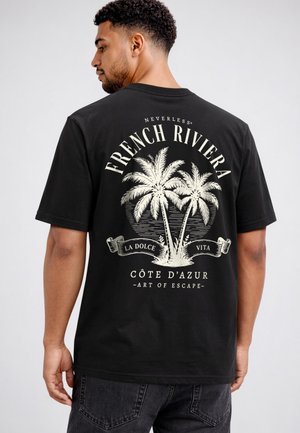Man wearing black t-shirt with white palm trees and text "French Riviera, La Dolce Vita, Côte d'Azur, Art of Escape" on the back.