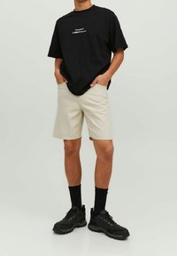 Muž v černém oversized tričku s bílým textem, béžových šortkách do kolen, černých ponožkách a černých teniskách, stojící s rukama v kapsách.