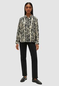Camicia a maniche lunghe color crema e nera con un design a foglia, abbinata a pantaloni neri a gamba dritta e scarpe slip-on nere.