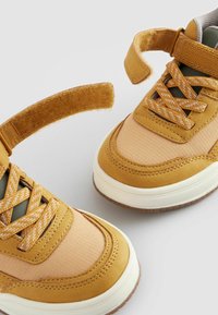 Geel en beige sneakers met een combinatie van stof en suède, voorzien van een klittenbandsluiting, gestructureerde veters en een rubberen zool.