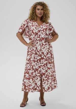 Mujer con cabello rizado, manos en las caderas, vestida con un vestido midi marrón óxido con estampado floral blanco y mangas abiertas en los hombros, sandalias negras de tacón.