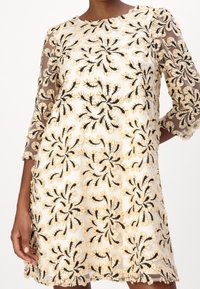 Broderad klänning med invecklade blommönster i beige och svart. Genomskinliga långärmar och en rundad halsringning med fransdetaljer vid fållen.