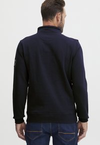 Pullover blu navy con colletto alto, maniche lunghe, polsini e orlo a costine. Presenta un logo circolare sulla spalla sinistra.