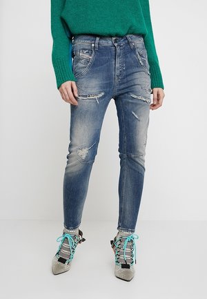 Osoba nosząca zielony sweter, sprane dopasowane niebieskie jeansy i buty z ostrym noskiem z turkusowymi sznurowadłami na jednolitym jasnym tle.