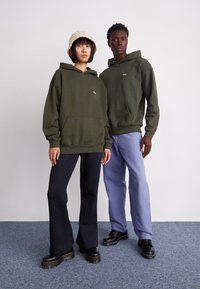 Des sweatshirts vert olive avec une poche avant, associés à un pantalon noir et bleu clair. La femme porte un chapeau seau beige ; tous deux portent des chaussures noires.
