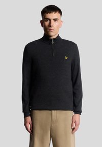 Svart zip-up-tröja med hög krage. Har en texturerad tyg och en liten gul logotyp på vänster bröst. Långa ärmar, åtsittande design.