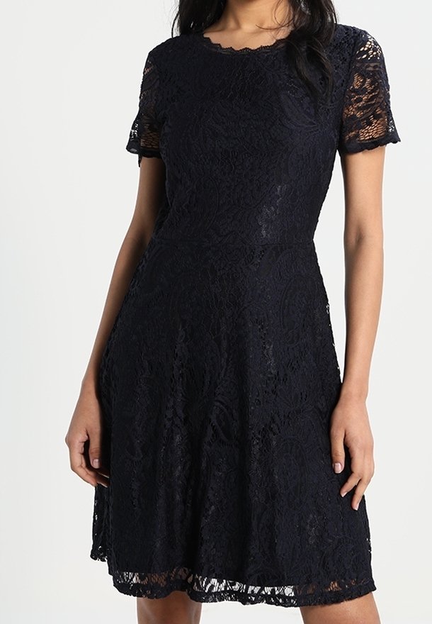 Femme portant une robe noire en dentelle jusqu'aux genoux avec manches courtes et col rond, debout devant un fond uni.