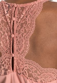 Spetsdetalj på baksidan av ett plagg i blommig ljusrosa färg, med två tygklädda knappar längs en nyckelhålsöppning.