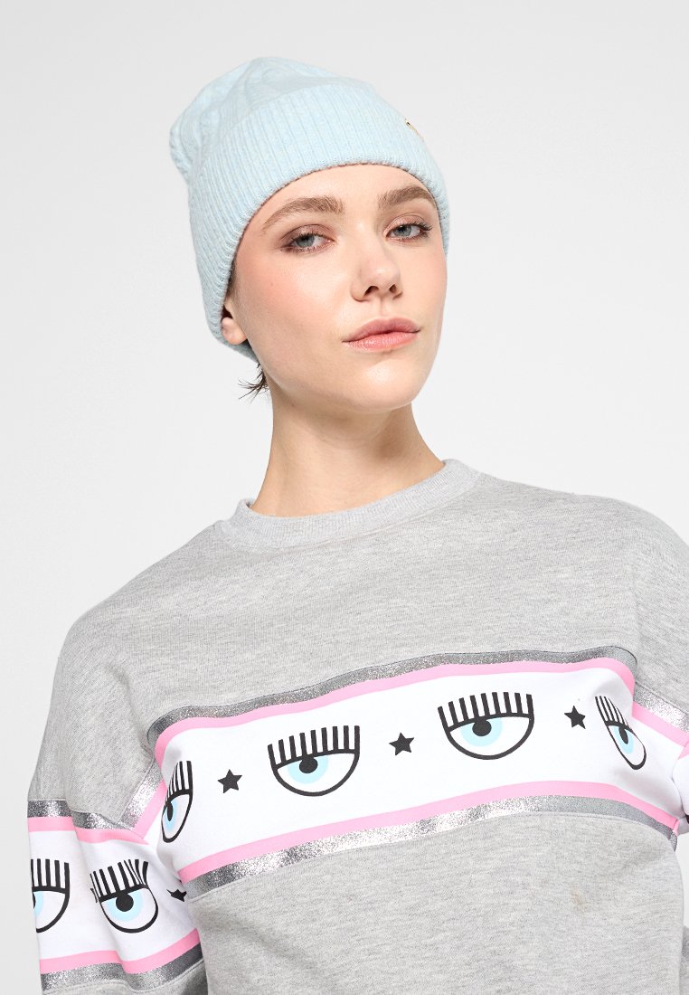 Hellblauer Strickmütze und grauer Sweatshirt mit einem auffälligen grafischen Design, das Augen, pinke Streifen und silberne Akzente zeigt.