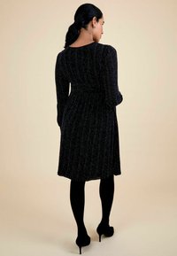 Vestido negro con manga larga, tejido texturizado que incluye hilos metálicos, cintura ceñida y una falda fluida. Lleva puesto con medias negras y tacones.